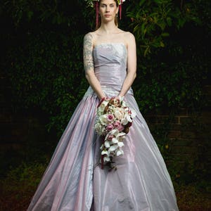 K&ouml;nnte beinhalten: Eine Braut in einem helllilafarbenen Brautkleid mit einer Blumenkrone und einem Strau&szlig; wei&szlig;er und rosa Blumen.