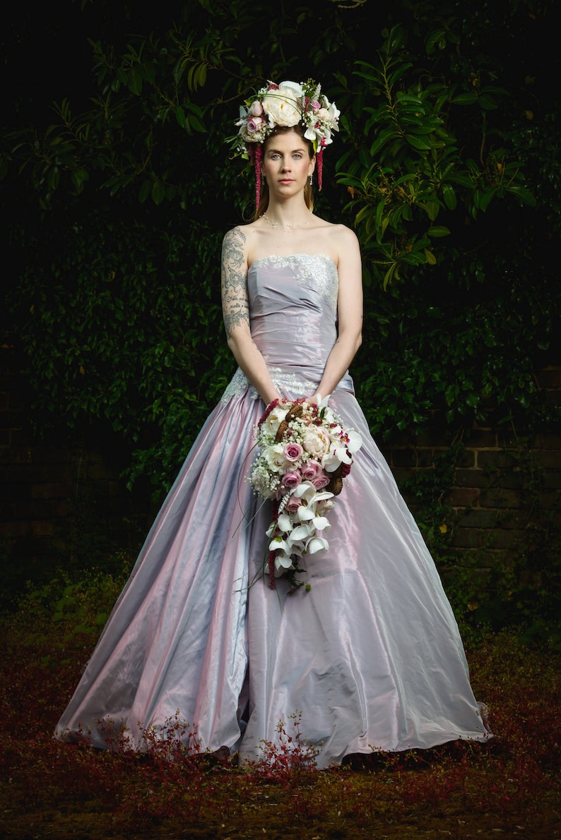 K&ouml;nnte beinhalten: Eine Braut in einem helllilafarbenen Brautkleid mit einer Blumenkrone und einem Strau&szlig; wei&szlig;er und rosa Blumen.