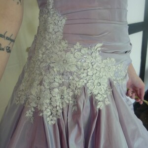 K&ouml;nnte beinhalten: Ein lavendelfarbenes Kleid mit einem wei&szlig;en Spitzenbesatz. Das Kleid hat ein tailliertes Oberteil und einen flie&szlig;enden Rock.