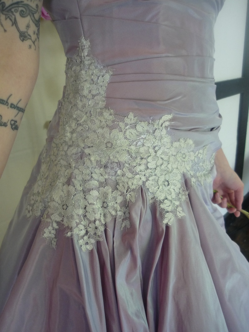 K&ouml;nnte beinhalten: Ein lavendelfarbenes Kleid mit einem wei&szlig;en Spitzenbesatz. Das Kleid hat ein tailliertes Oberteil und einen flie&szlig;enden Rock.