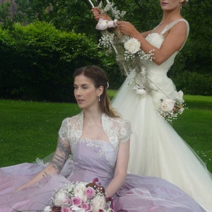 K&ouml;nnte beinhalten: Zwei Frauen in Hochzeitskleidung. Eine Frau tr&auml;gt ein wei&szlig;es Hochzeitskleid mit einem Blumenstrau&szlig;. Die andere Frau tr&auml;gt ein lavendelfarbenes Kleid mit einem wei&szlig;en und lavendelfarbenen Blumenstrau&szlig;.