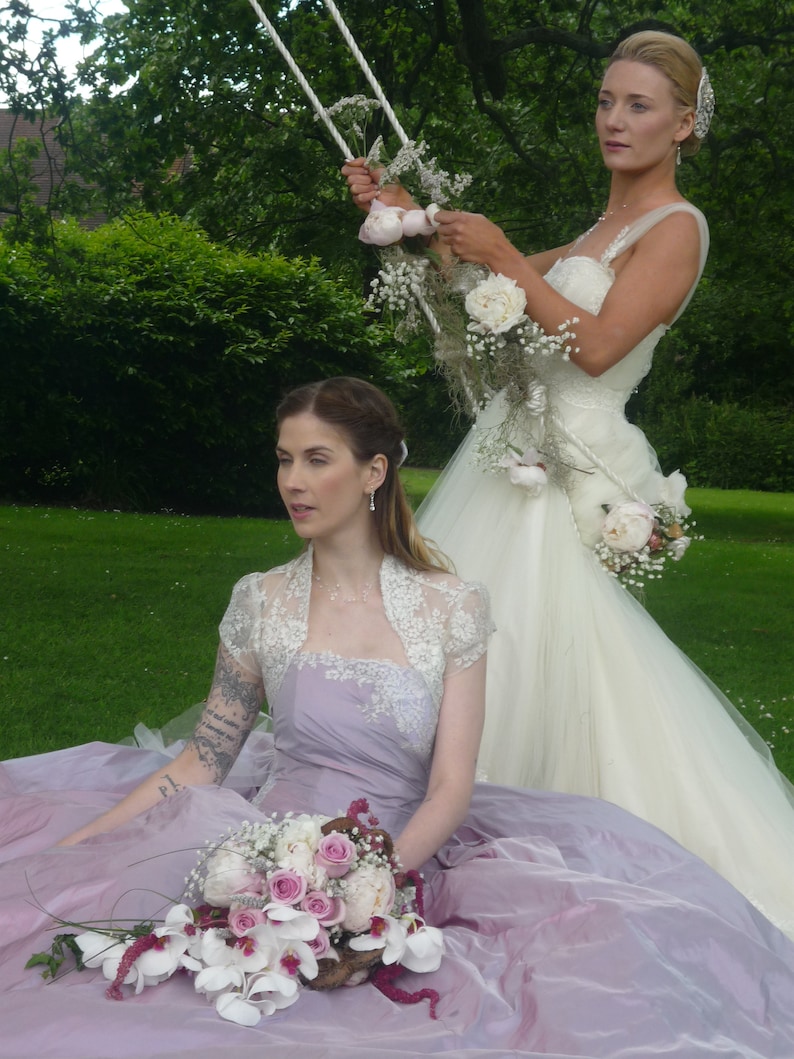 K&ouml;nnte beinhalten: Zwei Frauen in Hochzeitskleidung. Eine Frau tr&auml;gt ein wei&szlig;es Hochzeitskleid mit einem Blumenstrau&szlig;. Die andere Frau tr&auml;gt ein lavendelfarbenes Kleid mit einem wei&szlig;en und lavendelfarbenen Blumenstrau&szlig;.