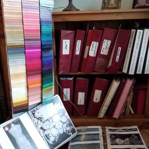 Puede incluir: Una estantería de madera con carpetas rojas etiquetadas con diferentes tipos de telas, incluyendo "Encaje", "Motivos", "Telas bordadas" y "Bennett Silks". La estantería está llena de muestras de telas y libros de muestras.