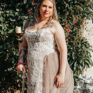 Könnte beinhalten: Ein trägerloses Brautkleid mit aufwendigen weißen Spitzendetails am Oberteil und einem fließenden, hellbraunen Tüllrock. Die Braut trägt ein silbernes Diadem und hält einen transparenten Gehstock mit rosa Akzenten. Der Hintergrund zeigt grünes Laub und rosa Blüten.