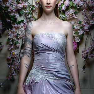 K&ouml;nnte beinhalten: Eine Frau in einem tr&auml;gerlosen Lavendelkleid mit Spitzendetails. Sie steht vor einer Wand, die mit rosa Blumen und Gr&uuml;npflanzen dekoriert ist.