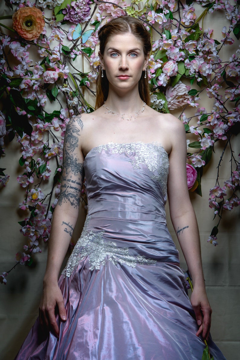 K&ouml;nnte beinhalten: Eine Frau in einem tr&auml;gerlosen Lavendelkleid mit Spitzendetails. Sie steht vor einer Wand, die mit rosa Blumen und Gr&uuml;npflanzen dekoriert ist.