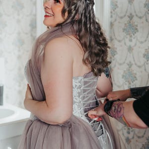 Könnte beinhalten: Ein lavendelfarbenes und weißes Korsettkleid mit Tüllrock wird geschnürt. Die Person, die das Kleid trägt, hat welliges, hellbraunes Haar und lächelt. Der Hintergrund zeigt florale Tapeten und ein Fenster.