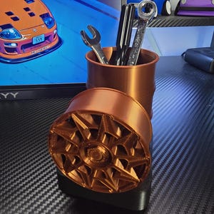 May include: A copper-colored desk organizer with a cylindrical pen holder and a decorative wheel-shaped base. The pen holder contains pens and wrenches. The wheel base is resting on a black base. The background includes a computer monitor and toy cars.