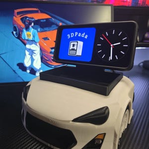 May include: A white 3D-printed car model with black accents, holding a black screen displaying a clock and the text "3DPada". The screen has a blue background with a 3D printer icon. A computer monitor with a car graphic is in the background.