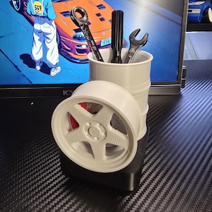 May include: A white, 3D-printed car wheel and a white barrel-shaped container holding tools. The tools include a wrench and a screwdriver. The wheel is positioned in front of the container. The background features a computer monitor with a car-themed graphic.