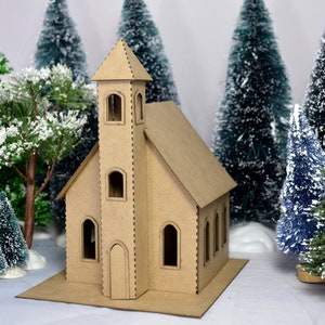 May include: A cardboard model of a church with a tall steeple, surrounded by miniature snow-covered pine trees. The church is made of brown cardboard and has a simple design.