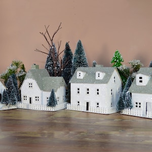 May include: A miniature white paper village with a church, houses, and trees. The houses have white picket fences and the trees are covered in glitter.
