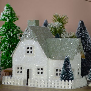May include: A white cottage with a green roof and white picket fence, surrounded by miniature evergreen trees covered in snow. The cottage has windows and a door, and the roof is covered in glitter.