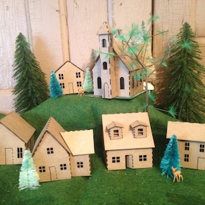 May include: A miniature cardboard village with a church, houses, and trees. The houses are brown and have white trim. The trees are green and have white snow on them. The village is set on a green surface.