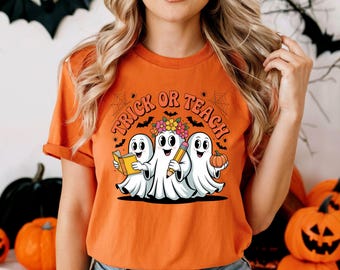 May include: Orange t-shirt featuring the words "Trick or Teach" and a graphic of three cartoon ghosts. One ghost holds a book, another a pencil, and the third a pumpkin. A Halloween-themed design.