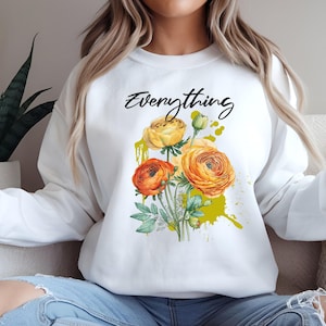 May include: White sweatshirt featuring the word "Everything" in black script above a floral design. The floral design includes orange and yellow flowers with green leaves and a yellow paint splatter.