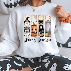 Peut inclure: Sweat-shirt blanc avec un motif d'Halloween. Le motif comprend un fantôme, une citrouille, un chat noir et une pierre tombale, avec les mots "Spooky Season" en dessous. Le sweat-shirt est porté.