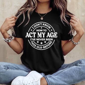 May include: Black t-shirt with a white circular graphic that reads "I DON'T KNOW HOW TO ACT MY AGE I'VE NEVER BEEN THIS OLD BEFORE". The shirt is worn by a person sitting down, wearing blue jeans and white trainers.