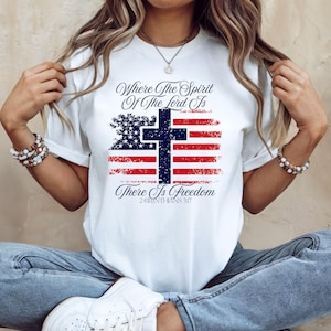以下が含まれることがあります： 愛国的なデザインの白いTシャツ。グラフィックには、様式化されたアメリカ国旗の上に十字架が重ねられ、「Where The Spirit Of The Lord Is, There Is Freedom」というテキストと聖書の詩2コリント3:17が描かれています。