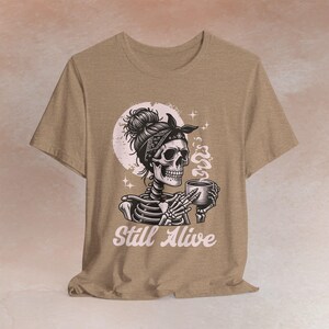 May include: A black t-shirt with a pink and black skull illustration wearing a bandana and holding a cup. The skull has a bun and is surrounded by a pink moon. The text 'Still Alive' is printed in pink below the illustration.