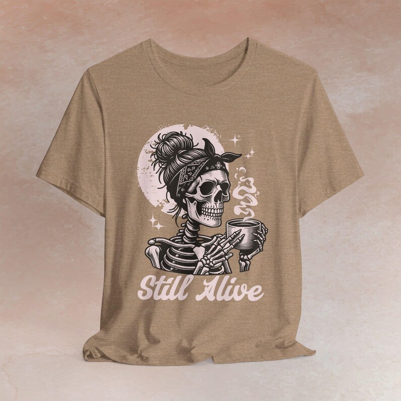 May include: A black t-shirt with a pink and black skull illustration wearing a bandana and holding a cup. The skull has a bun and is surrounded by a pink moon. The text 'Still Alive' is printed in pink below the illustration.
