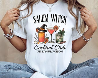 May include: White t-shirt with the text "Salem Witch Cocktail Club" and the phrase "Pick Your Poison". The graphic features three cocktail glasses with orange slices, a witch hat, and other decorative elements.