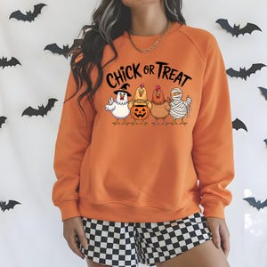 May include: An orange sweatshirt featuring the phrase "Chick or Treat" and a graphic of four chickens in Halloween costumes. The chickens are dressed as a witch, holding a pumpkin, and as a mummy. Black bat decorations are in the background.