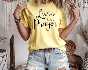 Peut inclure: Un t-shirt jaune avec l'inscription "Livin on a Prayer" en écriture noire. Le t-shirt est associé à un short en jean bleu clair et une ceinture marron. Le modèle porte des bracelets et un collier.