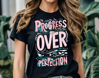 May include: Black t-shirt featuring the phrase "PROGRESS OVER PERFECTION" in pink and white. The design incorporates floral and abstract patterns in teal, green, and white. A small daisy is above the word "OVER".