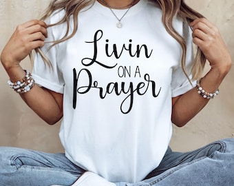 May include: White t-shirt with the phrase "Livin on a Prayer" in black script. The shirt is a classic crew neck style. The model is wearing blue jeans and white sneakers.