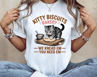 May include: White t-shirt with a graphic of a gray tabby cat kneading dough on a wooden board. The text "KITTY BISCUITS BAKERY" is above the cat, and "WE KNEAD EM - YOU NEED EM" is below. The design is in brown and black.