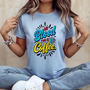 May include: Pale blue t-shirt featuring the phrase "My Blood Type Is Coffee" in a vibrant design. The text is in blue, yellow, and red, with a coffee cup illustration. A casual wear item.