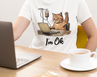 May include: White t-shirt featuring a cartoon cat working on a laptop with a coffee IV drip. The cat is orange and brown, with one paw raised. The text "I'm Ok" is printed below the cat.