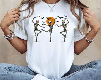 May include: White t-shirt with a Halloween-themed design. The graphic depicts three dancing skeletons, an orange sun, and bats. The skeletons are in ballet poses, creating a spooky yet playful image.