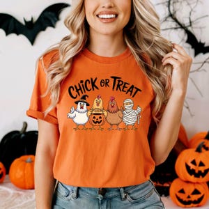 May include: Orange t-shirt featuring the text "Chick or Treat" and a graphic of four cartoon chickens in Halloween costumes. The chickens are dressed as a witch, a pumpkin, and a mummy.