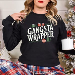 Puede incluir: Sudadera negra con el texto "GANSTA WRAPPER" en blanco y verde. El diseño incluye cajas de regalo, un árbol de Navidad y un adorno. La persona lleva un pantalón de pijama a cuadros y sostiene una taza blanca.