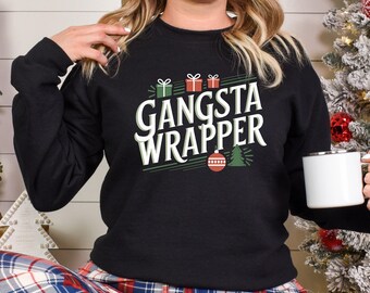 Pode incluir: Camisola preta com o texto "GANSTA WRAPPER" em branco e verde. O design inclui caixas de presente, uma árvore de Natal e um enfeite. A pessoa está vestindo calças de pijama xadrez e segurando uma caneca branca.