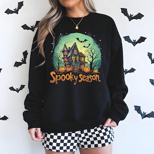 Peut inclure: Sweat-shirt noir avec un motif coloré d'Halloween. Le motif représente une maison hantée, des citrouilles, des chauves-souris et le texte "Spooky Season."