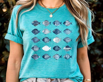 May include: Teal t-shirt featuring a pattern of stylized fish in various shades of blue and white. The fish designs include polka dots, stripes, and other geometric patterns. The shirt has rolled-up sleeves and a crew neck.