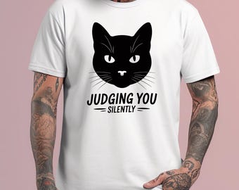May include: White t-shirt featuring a black cat face graphic with the text "JUDGING YOU - SILENTLY" in black. The cat graphic has white eyes and whiskers. The shirt is made of a soft, comfortable material.