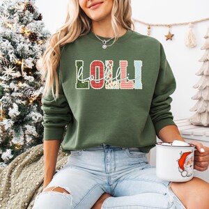 Może przedstawiać: Oliwkowo-zielona bluza z napisem "Lolli" w dekoracyjnym, wielokolorowym wzorze. Litery wypełnione są elementami kwiatowymi i wzorzystymi. W dłoni trzymany jest biały kubek z ilustracją Świętego Mikołaja.