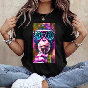 May include: Black t-shirt featuring a vibrant, pop art style image of a monkey wearing round glasses and licking a lollipop. The monkey's fur is a mix of pink, purple, and blue, with colorful accents in the background.
