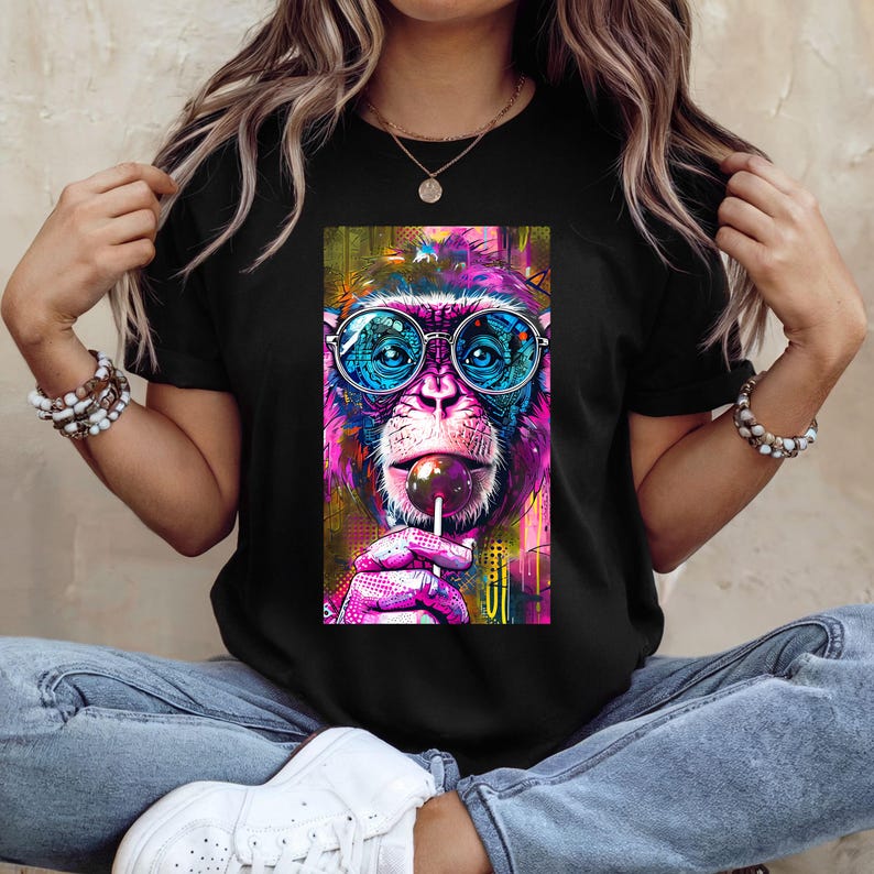 May include: Black t-shirt featuring a vibrant, pop art style image of a monkey wearing round glasses and licking a lollipop. The monkey's fur is a mix of pink, purple, and blue, with colorful accents in the background.