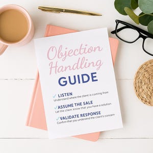 May include: A pink notebook with a white sheet of paper that reads "Objection Handling GUIDE" in pink and blue. A gold pen, a pink mug with coffee, black glasses, and a woven coaster are also on the white surface.