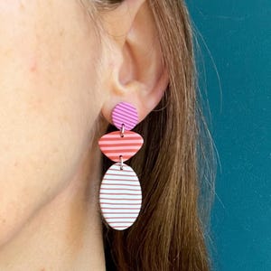 Peut inclure: Une boucle d'oreille pendante composée de trois formes géométriques superposées : un petit cercle rayé violet, un ovale rayé rouge moyen et un grand ovale blanc à fines rayures horizontales rouges, reliés par de petits anneaux en métal argenté.