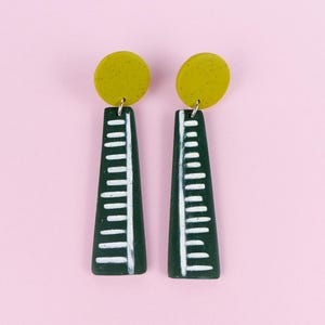 Peut inclure: Une paire de boucles d'oreilles faites à la main. Chaque boucle d'oreille présente un dessus circulaire de couleur chartreuse et un pendentif allongé vert foncé. Le pendentif a un motif en forme d'échelle blanche. Les boucles d'oreilles sont présentées sur un fond rose.
