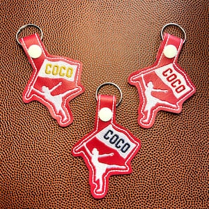 May include: Three red keychains with a white embroidered dancer design and the word "COCO" in a banner. Each keychain has a silver ring and a white button closure. The keychains are on a brown textured surface.