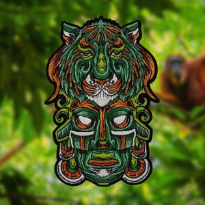 Op de afbeelding: Geborduurde patch met een groen en oranje tijgerontwerp boven een groen masker. De patch heeft een zwarte rand en gedetailleerde kenmerken. De achtergrond is een wazige afbeelding van groen gebladerte.