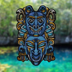 Pode incluir: Patch bordado com um design de máscara estilizada em camadas em tons de azul, dourado e branco. O patch retrata dois rostos, um sobre o outro, com detalhes intrincados e uma estética tribal. O fundo está desfocado.