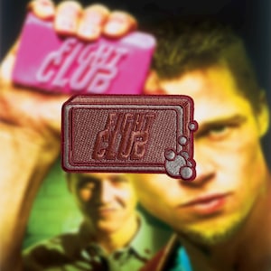 以下が含まれることがあります： 「FIGHT CLUB」の白い文字が入った、マルーン色の刺繍パッチ。右側に泡のデザインがあります。長方形のパッチにはステッチの縁取りがあります。同じテキストが入ったピンク色の石鹸が背景にあります。